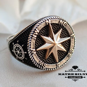 May include: A silver ring with a compass design featuring a gold-colored star. The compass has the directions N, E, S, and W inscribed around the edge. The ring also has a small ship's wheel design on the side.
