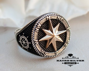 May include: A silver ring with a compass design featuring a gold-colored star. The compass has the directions N, E, S, and W inscribed around the edge. The ring also has a small ship's wheel design on the side.