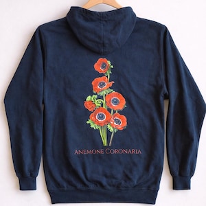 Puede incluir: Sudadera con capucha azul marino con un diseño floral de flores de anémona coronaria rojas con centros negros y tallos verdes. El texto "ANEMONE CORONARIA" está impreso debajo. La sudadera tiene capucha y mangas largas.