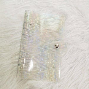 May include: A clear, iridescent notebook with a silver spiral binding and a snap closure. The notebook has a textured, holographic pattern that reflects light. The cover is made of a flexible, transparent material. The notebook is closed.