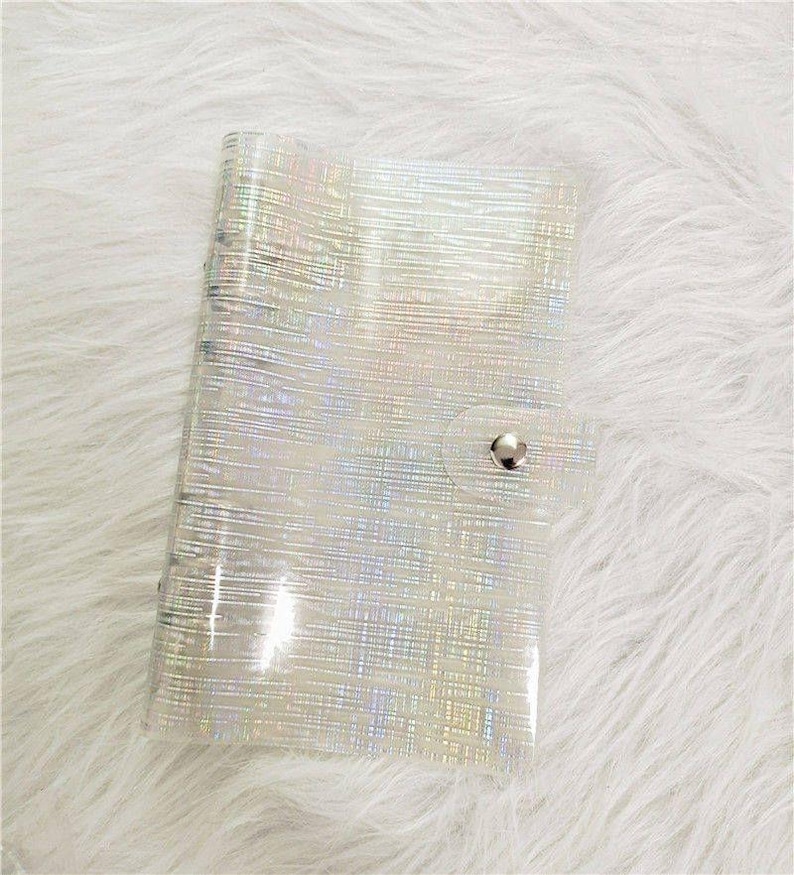 May include: A clear, iridescent notebook with a silver spiral binding and a snap closure. The notebook has a textured, holographic pattern that reflects light. The cover is made of a flexible, transparent material. The notebook is closed.