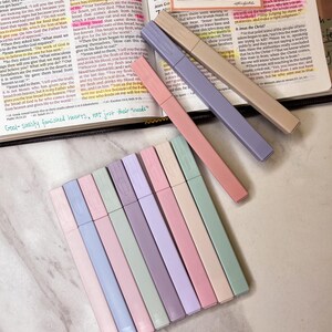 May include: An open book with highlighted text and colorful highlighters. The highlighters are in shades of pink, purple, beige, and green. The text on the page is partially visible.