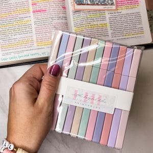 May include: A package of pastel-colored sticky notes is held in a hand, with a bracelet visible on the wrist. The notes are arranged in a neat stack, with a label that reads "Little Helpful Files PRINTABLES". The background includes an open book.