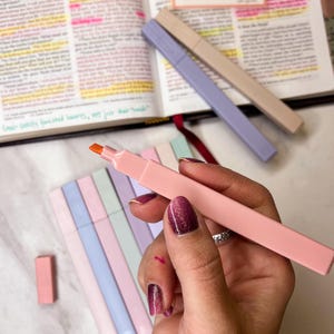 May include: A hand holding a pink highlighter with an orange tip, next to a set of pastel-colored highlighters. An open book with highlighted text and two other highlighters are in the background. The highlighters are for studying and note-taking.