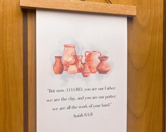 Może przedstawiać: Biała zawieszka ścienna z akwarelową ilustracją naczyń ceramicznych w kolorze terakoty. Poniżej grafiki znajduje się tekst: "But now, O LORD, you are our Father; we are the clay, and you are our potter; we are all the work of your hand." Isaiah 64:8. Wydruk jest zamocowany za pomocą dwóch jasnych drewnianych listewek.