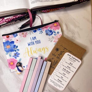 May include: A collection of Bible study supplies, including a floral print zippered pouch with the text "I AM WITH YOU Always", pastel highlighters, and a Bible highlighting color code card. An open Bible is visible in the background.