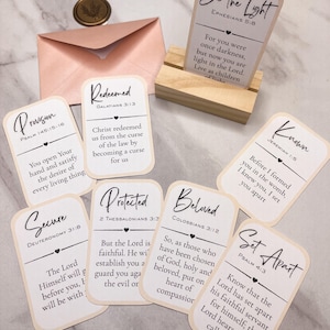 May include: A set of eight rectangular cards with rounded corners featuring black printed scripture verses and titles, a light pink envelope with a gold wax seal, and a small rectangular wooden stand holding one card that reads Be the Light.