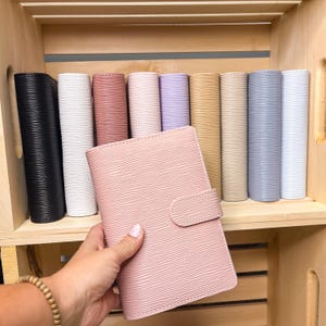 May include: A collection of rectangular planners with a textured, wavy horizontal line pattern. The planners are available in various solid colors including black, white, dusty rose, light pink, lavender, mustard yellow, beige, gray, and light blue.