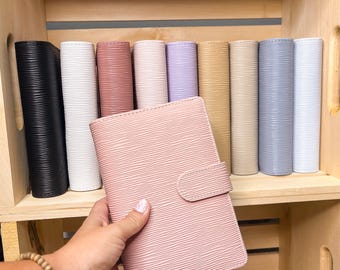 May include: A collection of rectangular planners with a textured, wavy horizontal line pattern. The planners are available in various solid colors including black, white, dusty rose, light pink, lavender, mustard yellow, beige, gray, and light blue.