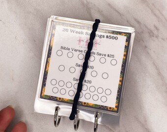 Może przedstawiać: Przezroczysty akrylowy tracker oszczędności z czarnym sznurkiem. Wkładka z nadrukiem "26 Week Savings $500" i "Bible Verse Cards Save $25". Wkładka ma obramowanie ze słoneczników i kółka do śledzenia oszczędności.