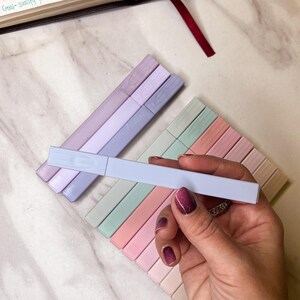 May include: A hand holding a light blue highlighter pen, with a set of pastel-colored highlighters in the background. The highlighters are arranged in a rectangular pattern, with colors including pink, green, and lavender. A book is open in the background.