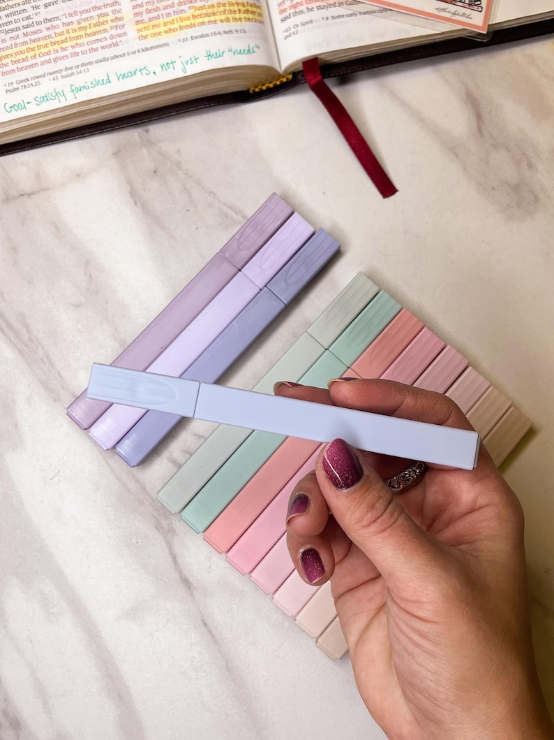 May include: A hand holding a light blue highlighter pen, with a set of pastel-colored highlighters in the background. The highlighters are arranged in a rectangular pattern, with colors including pink, green, and lavender. A book is open in the background.