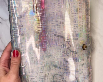 May include: A clear, iridescent planner with a grid pattern, held by a hand with red nail polish. Two pens are clipped to the top. The planner has a snap closure and handwritten text.