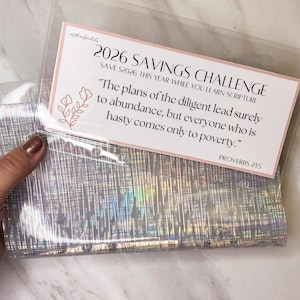 May include: A clear plastic savings challenge envelope with a pink and white card. The card reads "2026 Savings Challenge" and includes a Bible verse. The envelope has a holographic, iridescent design.