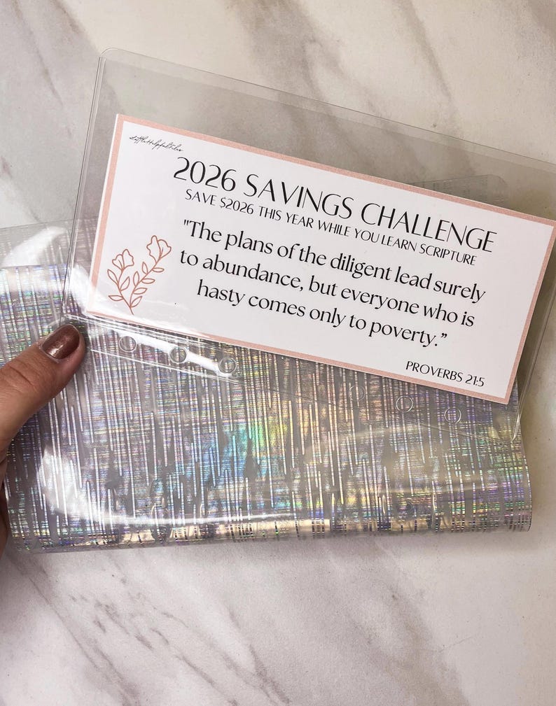 May include: A clear plastic savings challenge envelope with a pink and white card. The card reads "2026 Savings Challenge" and includes a Bible verse. The envelope has a holographic, iridescent design.
