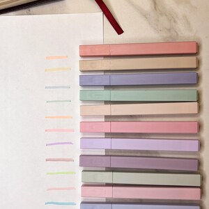 May include: A collection of pastel-colored highlighters arranged next to corresponding color swatches on a white surface. The highlighters are in shades of pink, beige, purple, green, and blue. A Bible with a red ribbon bookmark is visible in the background.