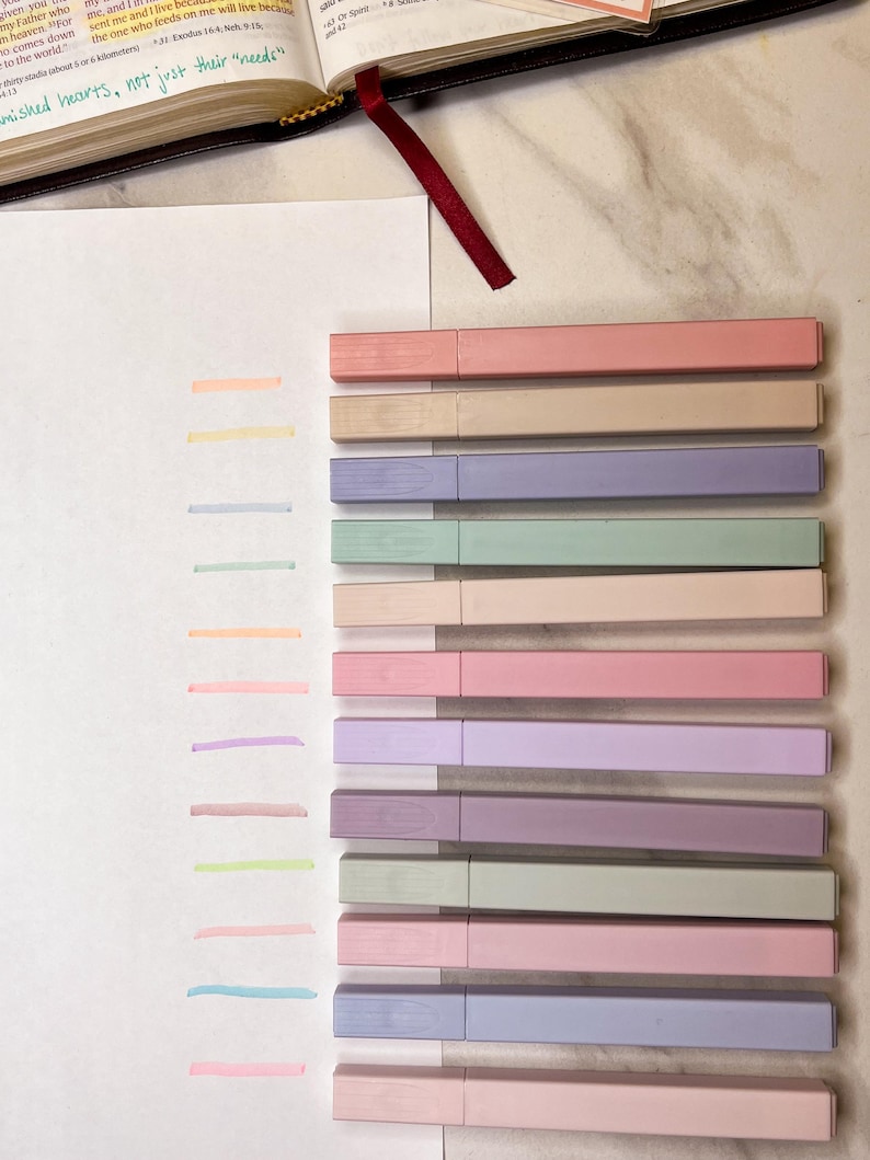 May include: A collection of pastel-colored highlighters arranged next to corresponding color swatches on a white surface. The highlighters are in shades of pink, beige, purple, green, and blue. A Bible with a red ribbon bookmark is visible in the background.