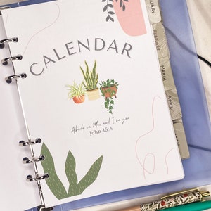 Könnte beinhalten: Ein Planer mit einer Titelseite "KALENDER" mit Illustrationen von Topfpflanzen. Die Seite ist auf der Januar-Registerkarte geöffnet. Zwei Stifte mit floralen und textlichen Details sind sichtbar. Der Text "Abide in Me, and I in you John 15:4" ist ebenfalls sichtbar.