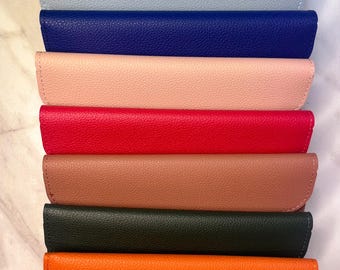 May include: A stack of eight rectangular wallets with a textured finish, arranged vertically. The colors from top to bottom are black, light blue, royal blue, light pink, red, mauve, dark green, and bright orange, each featuring a flap closure.