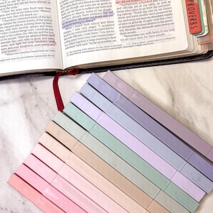 May include: Open Bible with highlighted text and colorful page markers. The open book displays text, with a red ribbon bookmark and several pastel-colored tabs, suggesting study or devotional use.