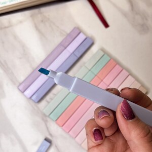 May include: A hand holding a white highlighter with a blue tip, with several pastel-colored highlighters in the background. The highlighters are arranged in rows of pink, peach, mint green, and lavender. A book with highlighted text is visible.