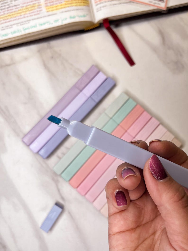 May include: A hand holding a white highlighter with a blue tip, with several pastel-colored highlighters in the background. The highlighters are arranged in rows of pink, peach, mint green, and lavender. A book with highlighted text is visible.