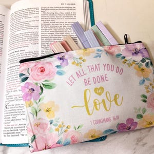 May include: A white pencil case with a floral design and the text "LET ALL THAT YOU DO BE DONE IN LOVE" in gold lettering. The case is open, revealing several colored highlighters. The case is resting on an open book.