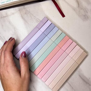 Peut inclure: Ensemble de surligneurs pastel, disposés en diagonale. Les couleurs incluent le lavande, le bleu, le vert, le rose et le beige. On aperçoit également une Bible avec un signet rouge et le texte "Goal-satisfy famished hearts, not just their needs."