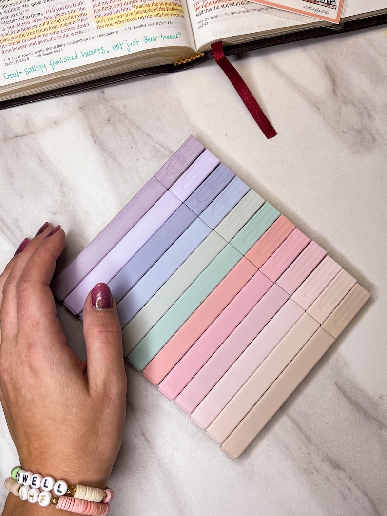 May include: A collection of pastel-colored highlighters arranged diagonally. The highlighters are in shades of lavender, blue, green, pink, and beige. The image also shows a Bible with a red ribbon bookmark and the text "Goal-satisfy famished hearts, not just their needs."