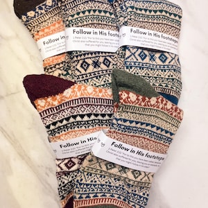 May include: A collection of patterned socks in various colors, including orange, blue, and green. Each sock features a unique geometric design and is wrapped with a label that reads "Follow in His footsteps".