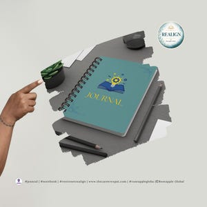 May include: A teal spiral-bound journal with the word "JOURNAL" in gold lettering. The cover features an open book graphic with a lightbulb and stars. Two pencils and a small potted plant are also visible.