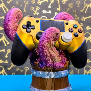 May include: A gold and black video game controller with blue accents, resting on a purple and pink tentacle-like structure. The controller is set on a brown barrel with silver trim, against a background of gold keys.
