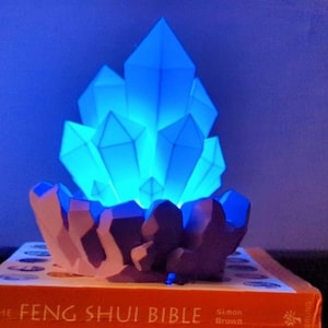 Puede incluir: Una luz nocturna con forma de cristal azul se encuentra sobre un libro titulado "The Feng Shui Bible" de Simon Brown. El cristal brilla y tiene una base morada.