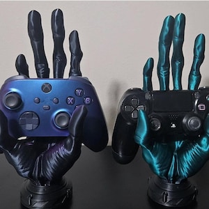 May include: Two 3D printed hands holding video game controllers. The hand on the left is black with a purple iridescent finish and is holding a blue Xbox controller. The hand on the right is teal green and is holding a black Playstation controller.