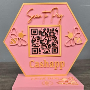 Może przedstawiać: Różowy i złoty sześciokątny znak z napisami "Scan to Pay" i "Cashapp". Znak zawiera kod QR i motywy pszczół. Znak znajduje się na różowej podstawie.