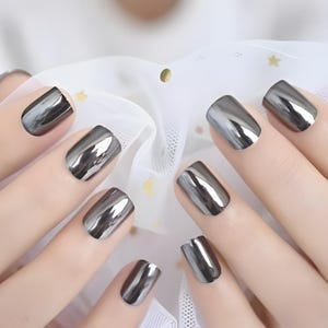 May include: Close-up of a set of metallic silver nail polish applied to short, square-shaped nails. The nails have a reflective, mirror-like finish, and are set against a soft, white background.