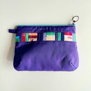 Puede incluir: Bolsa morada con cremallera y un diseño de patchwork colorido en la parte superior. El patchwork incluye rectángulos de varios colores y patrones, incluyendo telas florales y lisas. La cremallera es turquesa y el tirador es negro.