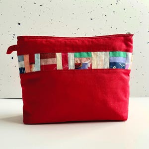 Puede incluir: Un estuche rojo con cremallera y una colorida banda de patchwork en la parte superior. El patchwork incluye varios retazos de tela en tonos rosa, azul, verde y crema. El estuche tiene un pequeño lazo de tela en el lateral.