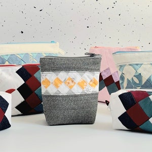 May include: A selection of handmade fabric pouches and zippered bags in various sizes. The bags feature patchwork designs in colours like pink, white, grey, blue, and burgundy. Some have diamond patterns, others striped or solid designs.