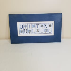May include: A framed cross-stitch with the word "QUILTING" spelled out in blue and white letters. Each letter is decorated with a small, silver charm related to quilting.