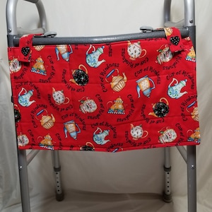 May include: Red fabric walker bag with a teacup and teapot pattern. The bag has the words "Cup of Kindness" repeated throughout the design. The bag is attached to a walker with two black clips.
