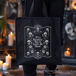 May include: Black tote bag with a white framed design featuring skulls and flowers, and the text "It's a Beautiful Day to Leave Me Alone." The bag is held by a person with tattoos. The words "House of Van Del" are written in cursive.