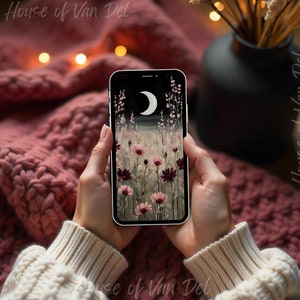May include: A smartphone displaying a digital painting of a field of pink and burgundy flowers with a crescent moon in the sky. The phone is held by a person's hands, with a pink knitted blanket in the background.
