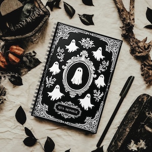May include: Black spiral bound notebook with a black and white design featuring ghosts and ornate scrollwork. The notebook has the name "Rita Monroe" printed on the front cover.