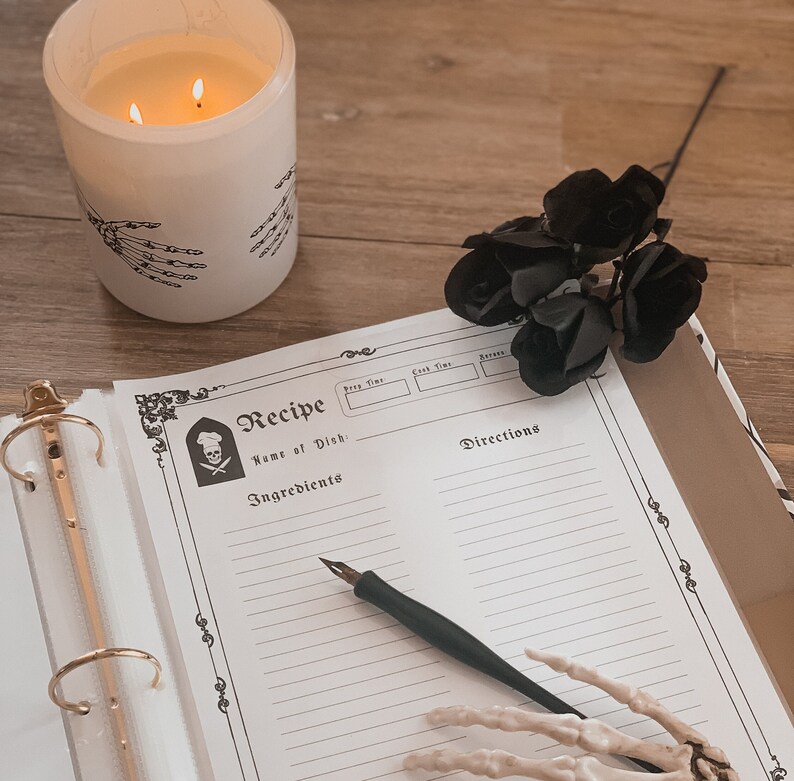 May include: A black and white recipe book with a skull and crossbones logo. The book is open to a blank page with lines for writing down ingredients and directions. A black quill pen and a skeleton hand are on the page.