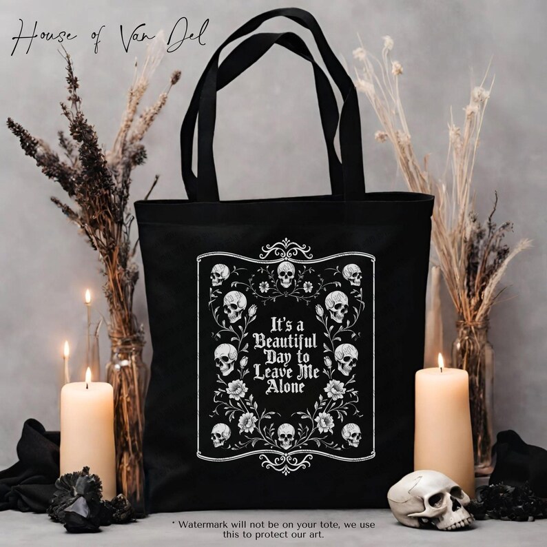 May include: Black tote bag with a white graphic featuring skulls, flowers, and the text "It's a Beautiful Day to Leave Me Alone." The bag has black handles and is surrounded by candles and dried plants.