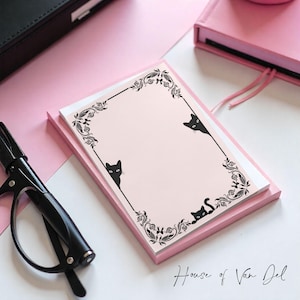May include: A pink notepad with a black floral border and black cat silhouettes. The notepad is stacked on top of a pink notebook. The text "House of Van Del" is written in cursive at the bottom right corner. A black pen and a black case are in the background.