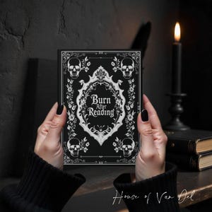 May include: A black book cover with the text "Burn After Reading" in a decorative frame, adorned with skulls and floral elements. The book is held against a dark background, with a lit candle and other books visible.