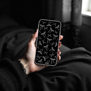 May include: A smartphone with a black screen displaying a pattern of gray bat and bow tie illustrations. The phone is held in a hand with dark nail polish, against a dark background. The phone case has a white border.