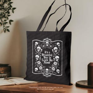 May include: Black tote bag with a white graphic design. The design features skulls, flowers, and the text "It's a Beautiful Day to Leave Me Alone" in a decorative frame. The bag has black handles.